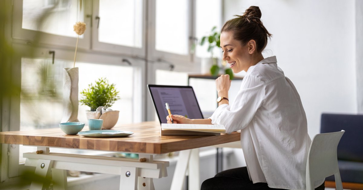 A woman sitting at her home office and smiling. She's writing something down and has her laptop turned on.