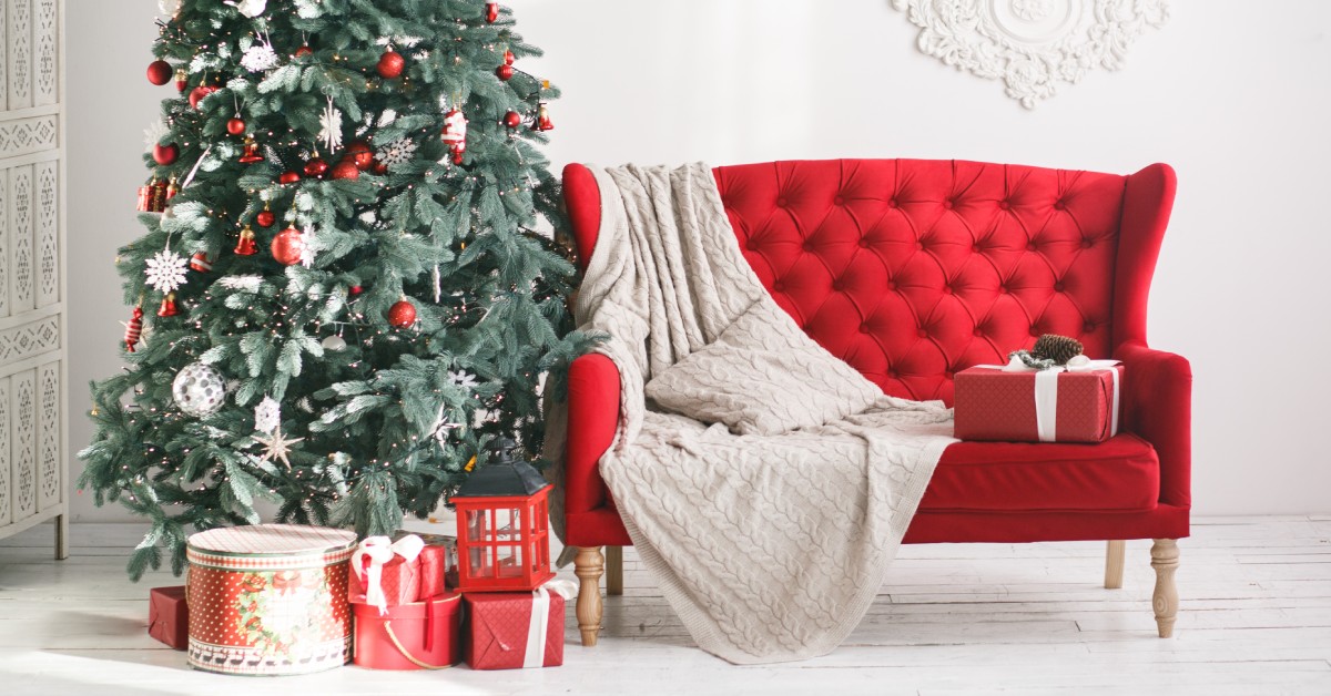 A red couch with a cover, a pillow, and a present sitting on it. It's next to a Christmas tree that has ornaments on it.