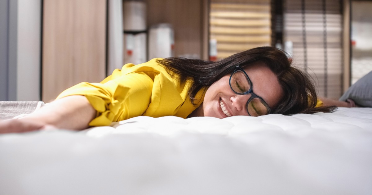 A person lying on a white mattress and smiling. She's wearing a yellow shirt and has glasses on.