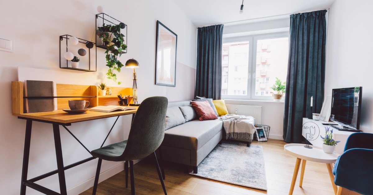 A living room with a desk, a couch, and a television. A sunny, curtained window and white walls fill the background.