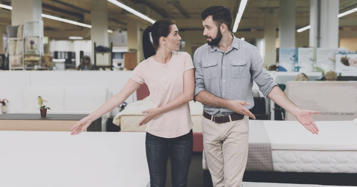 A man and a woman standing together in a furniture store. They're looking at each other and motioning toward different mattresses.