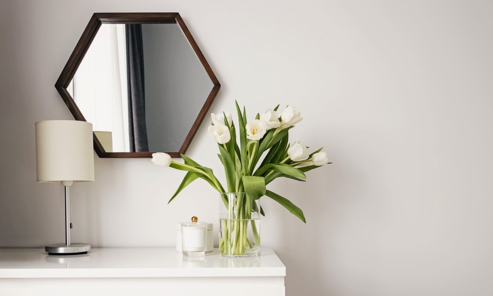 A close-up of a dresser with a lamp and vase of flowers on top of it. There is a mirror hanging on the wall above the dresser.